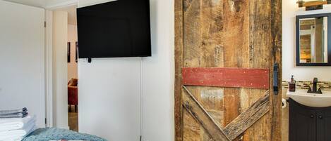 Une chambre à coucher avec un lit, un téléviseur fixé au mur, une porte en bois et une salle de bain avec un lavabo et un miroir.