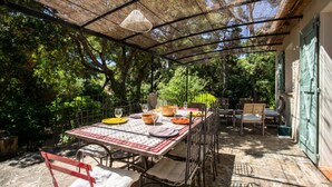 An outdoor patio with a table set for dining, surrounded by chairs and a shaded area with a woven ceiling. - House in Hyeres Near Almanarre Beach (Hyères)