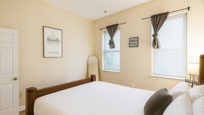 A bedroom with a bed, a door, a framed picture of a cityscape, and two windows with curtains. - Pet-Friendly Soulard Townhouse · 2BR/2BA · Parking + Rooftop Deck (St. Louis)