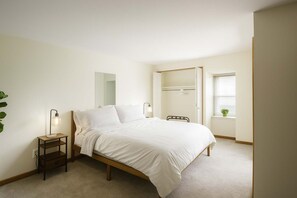Ein Schlafzimmer mit Bett, Nachttisch, Lampe und einem Fenster mit Pflanze.