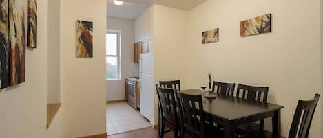 A dining area with a table and chairs, a kitchen in the background, and abstract art on the walls.