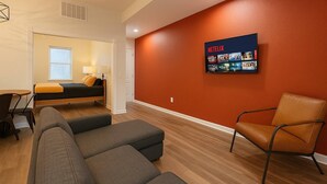 Une chambre d’hôtel moderne équipée d’un lit, d’un coin salon avec un canapé et des fauteuils, d’un téléviseur fixé au mur diffusant du contenu Netflix et d’une fenêtre avec des stores.