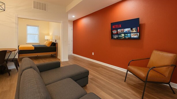 Une chambre d’hôtel moderne équipée d’un lit, d’un coin salon avec un canapé et des fauteuils, d’un téléviseur fixé au mur diffusant du contenu Netflix et d’une fenêtre avec des stores.