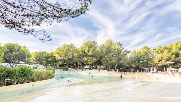 A water park with multiple pools and a sandy beach area, surrounded by trees and shaded by a large tree. - Apartment in Pont Royal With Water Park Fun (Mallemort)