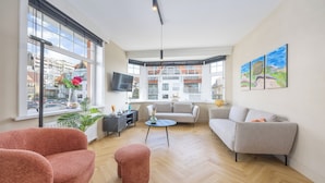 A spacious living room with large windows, a grey sofa, a pink armchair, and a small round table. - Holiday Home in Middelkerke Near Beach (Middelkerke)