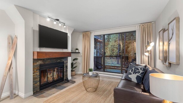 Ein modernes Wohnzimmer mit Kamin, Flachbildfernseher, einer Couch und einem großen Fenster mit Blick auf Bäume.