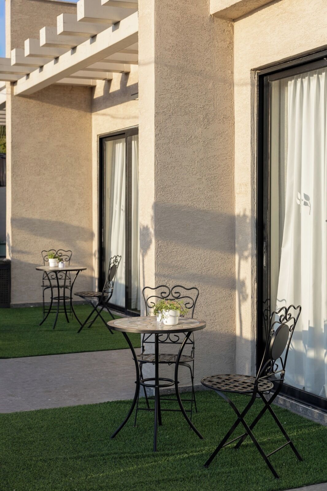 Un patio con dos mesas y sillas, una pequeña planta en maceta sobre cada mesa, y un edificio con fachada texturizada y ventanas amplias.