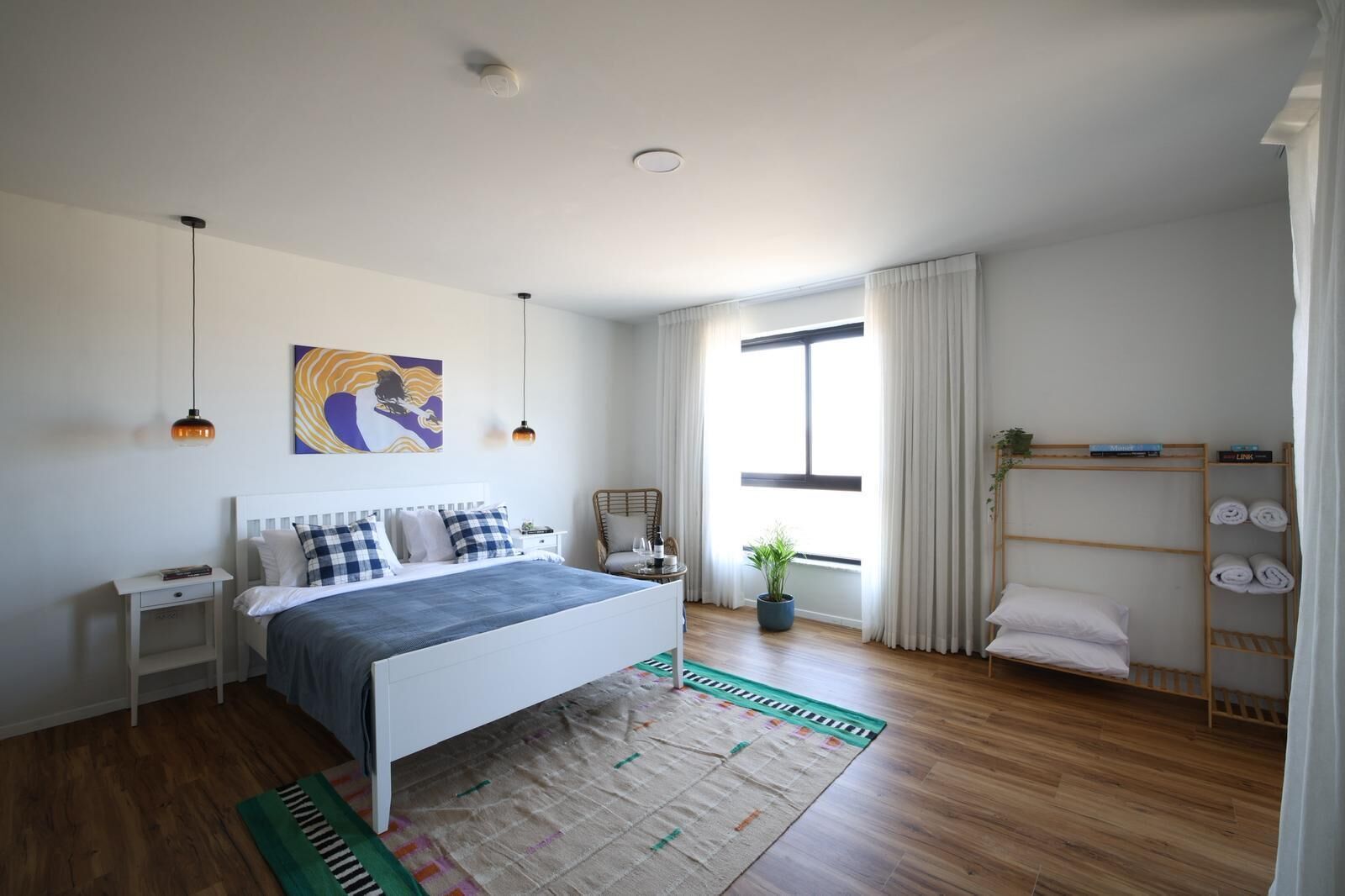 Une chambre moderne avec un lit, des tables de chevet, une fenêtre avec des rideaux, une plante en pot et une étagère avec des serviettes.