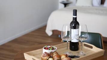 A wooden table with a tray containing croissants, a bottle of wine, and two glasses, set in a hotel room with a bed and a nightstand in the background.