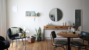 Un salon moderne avec une table de salle à manger ronde en bois, des chaises noires, un canapé noir, une petite table d’appoint avec un panier et une plante, un bureau en bois avec un miroir et une étagère avec des photos encadrées et une plante.