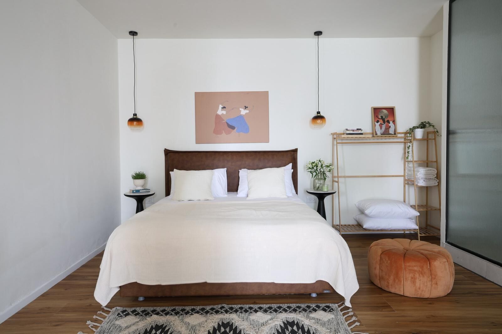 Ein Schlafzimmer mit einem großen Bett, einem hölzernen Kopfteil, einem Nachttisch mit einer Pflanze, einem kleinen Regal mit gefalteten Handtüchern und einer hängenden Deckenleuchte.