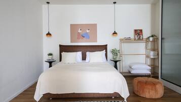 Ein Schlafzimmer mit einem großen Bett, einem hölzernen Kopfteil, einem Nachttisch mit einer Pflanze, einem kleinen Regal mit gefalteten Handtüchern und einer hängenden Deckenleuchte.
