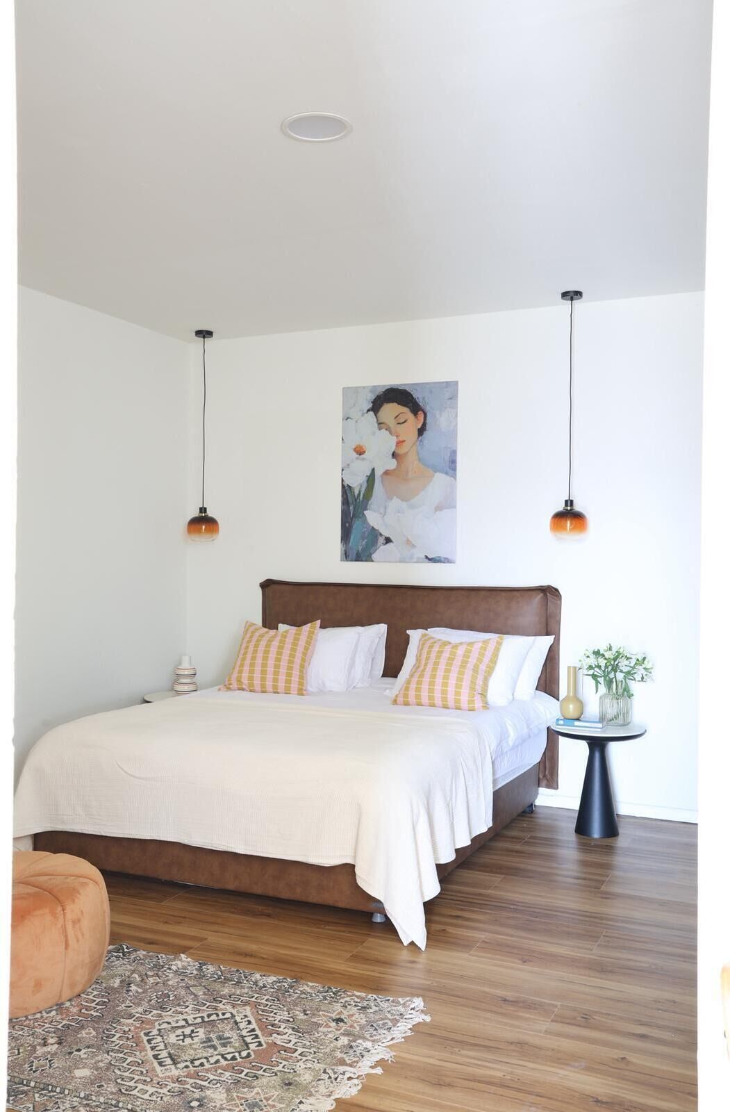 A bedroom with a large bed, two pendant lights, a painting on the wall, a small table with a vase, and a rug on the floor.