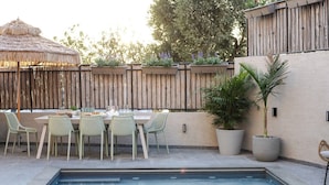 Un espace aménagé au bord de la piscine, avec une table dressée pour quatre personnes, entourée de chaises en osier, d’une clôture en bois et de plantes en pot.