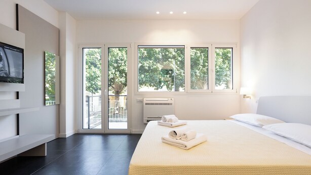 A modern hotel room with a large bed, a flat-screen TV, and a balcony with a view of trees. - Luxury Apartment Cefalù Giglio (Cefalù)