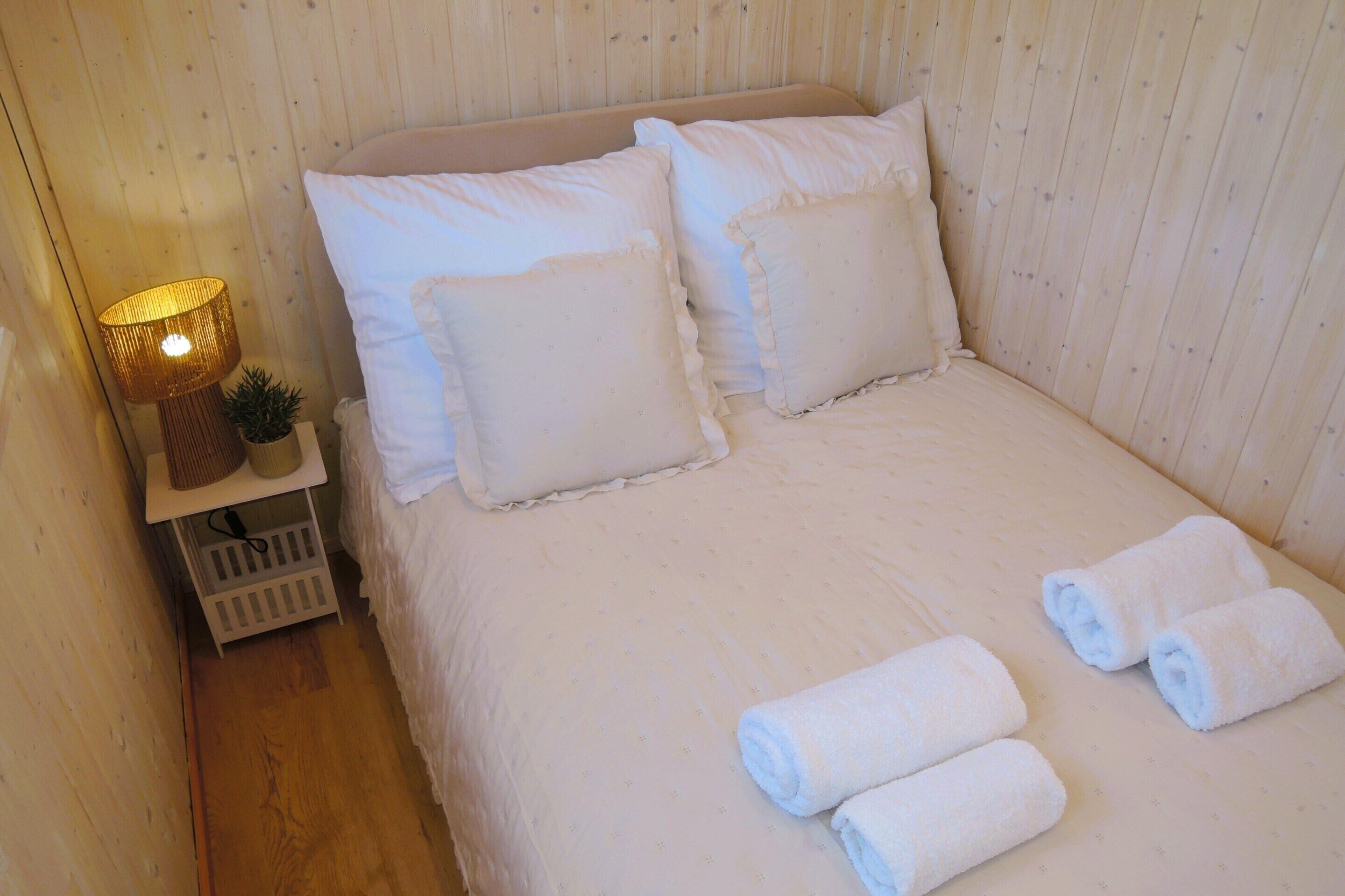 A neatly made bed with white linens and pillows, a bedside lamp, and two rolled towels on the floor.