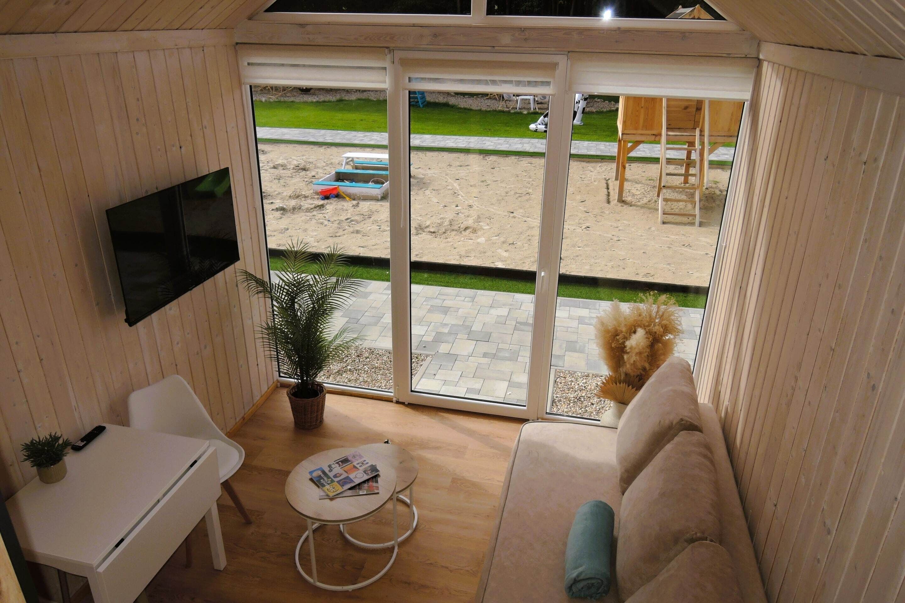 A room with a TV, a small table, a sofa, and a view of a sandy area with a wooden structure.
