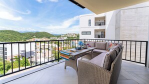Ein Balkon mit einer Rattan-Couch und einem Tisch, mit Blick auf eine Stadtlandschaft und grüne Hügel.