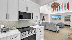 A modern kitchen with white cabinets, a microwave, a stove, and a toaster. There is a gray sofa and a television in the living area. - Chilli Fish 4 Bedroom Home (Anna Maria)