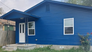 Une maison bleue avec une porte blanche et deux fenêtres, entourée d’une cour verdoyante.
