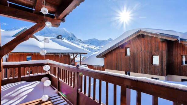 Een houten chalet met een balkon en sneeuwbedekte daken, gelegen tegen een bergachtige achtergrond.