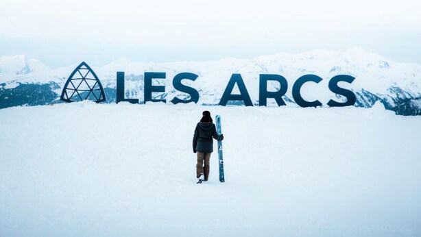 Una persona de pie frente a un letrero que dice "LES ARCS", con una montaña cubierta de nieve al fondo.