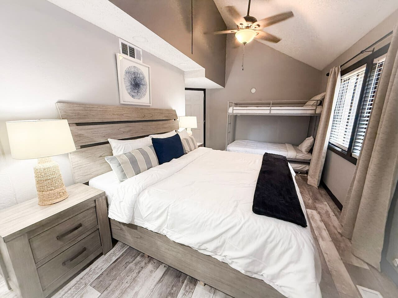 Un dormitorio con cama, mesitas de noche, ventilador de techo y ventana con cortinas.