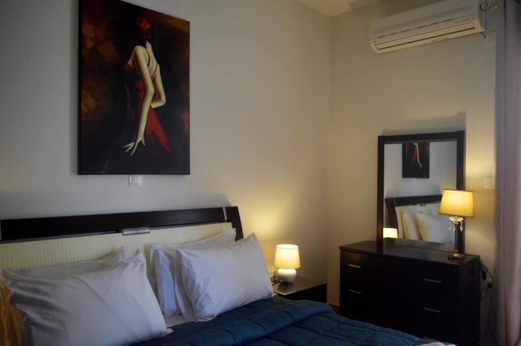 Una habitación de hotel con una cama, una mesita de noche con lámpara, un aparador, un cuadro en la pared y un aire acondicionado.