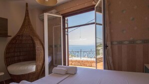 A bedroom with a bed, a wicker chair, and a balcony with a view. - Malvazios Villas (East Mani)