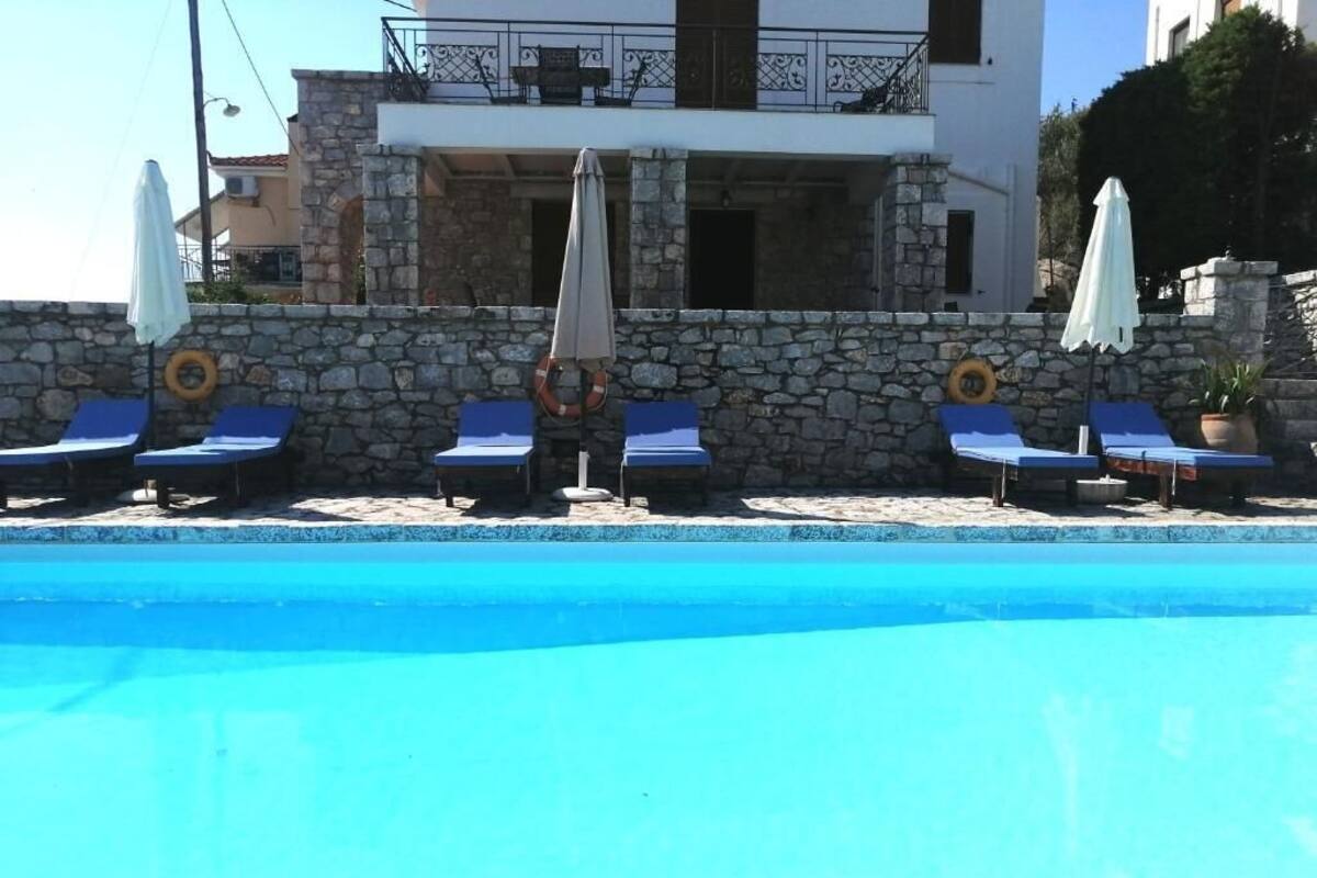 Un espace piscine avec des chaises longues et des parasols, un mur en pierre et un bâtiment avec des balcons en arrière-plan.