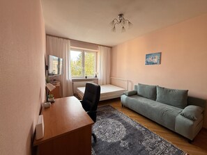 Ein kleiner, gut beleuchteter Raum mit einem Bett, einem Schreibtisch, einem Stuhl, einer Couch, einem Fenster mit Vorhängen und einem Bild an der Wand.