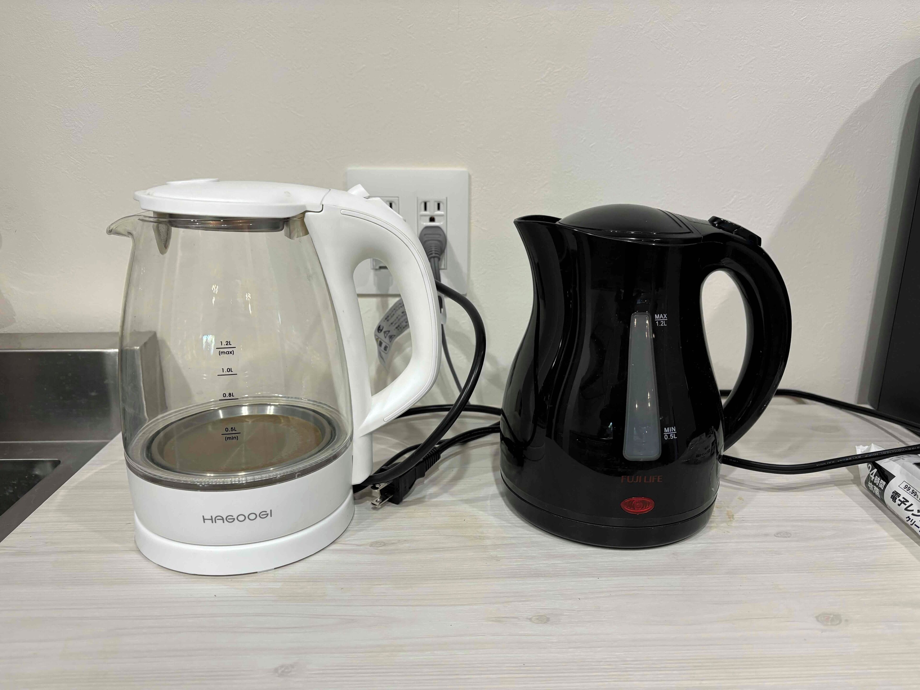 Une bouilloire électrique blanche de la marque HAGOOGI et une cafetière électrique noire de la marque FUJILIFE, posées sur une surface en bois.