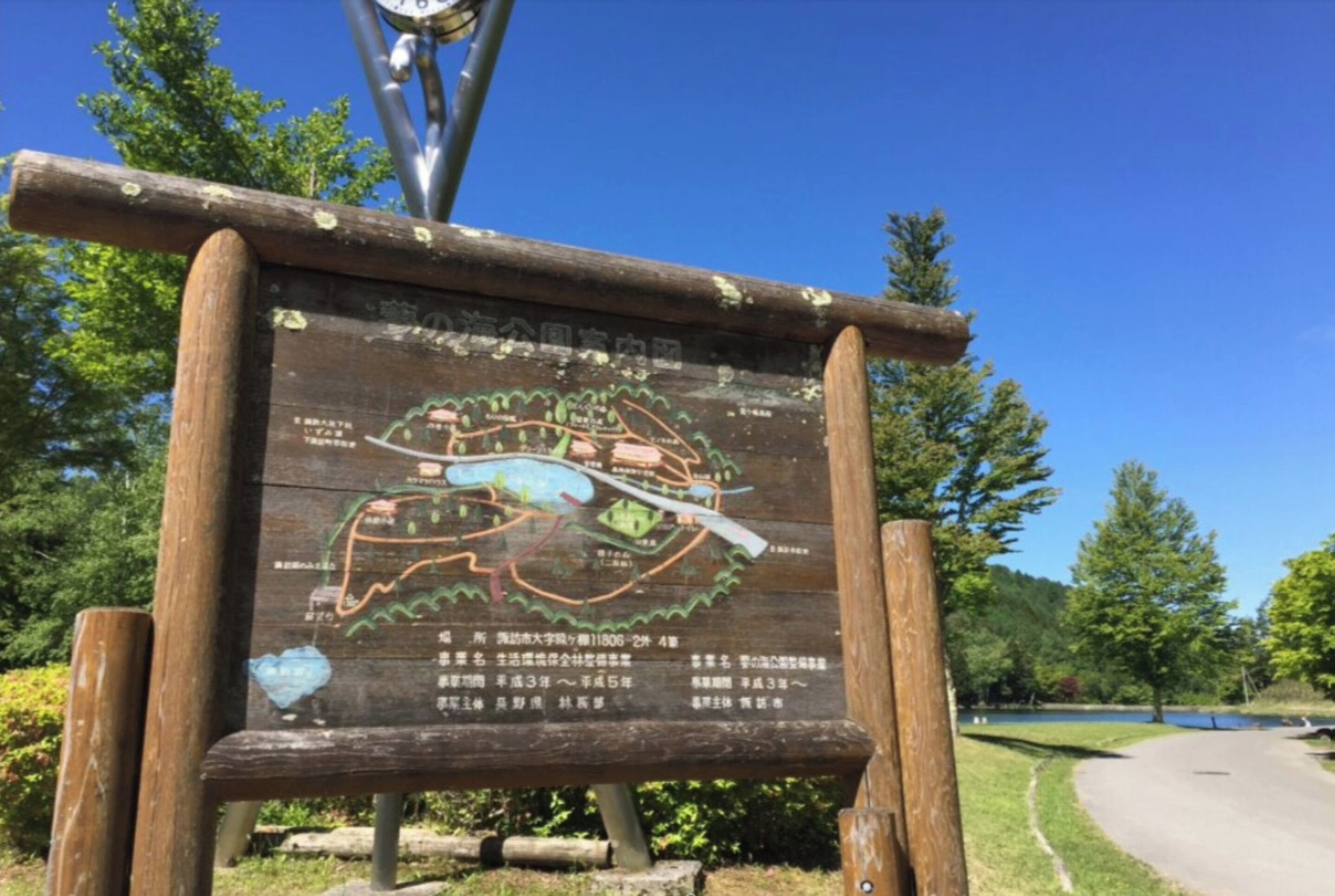 A wooden sign with a map and text, surrounded by trees and a clear sky.