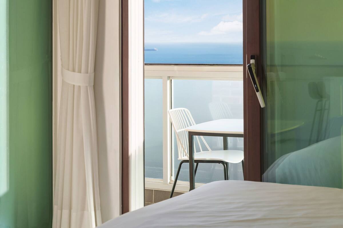 Une chambre d’hôtel avec un lit, une fenêtre avec des rideaux, un balcon avec une table et des chaises, et une vue sur la mer.