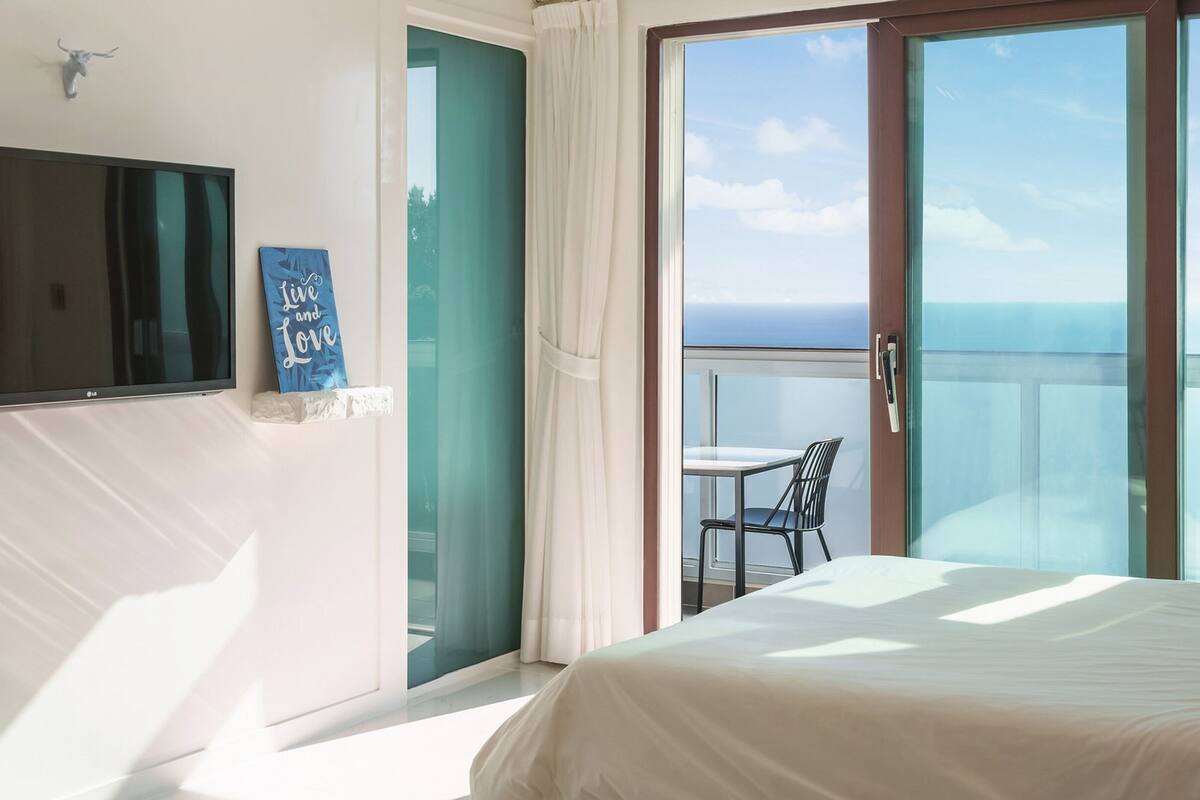 Une chambre à coucher comprenant un lit, une télévision et une porte-fenêtre coulissante donnant sur un balcon équipé d’une chaise et d’une table.