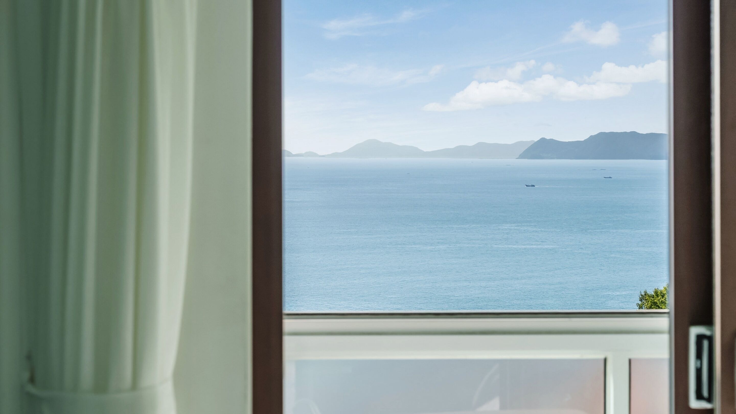 A view through a window with a curtain, overlooking a calm sea with distant mountains and a clear sky.