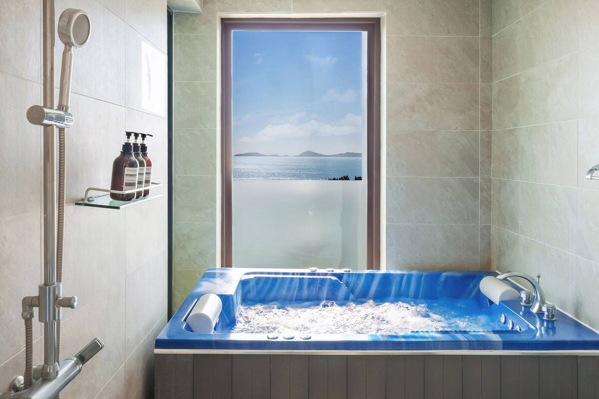 Une salle de bain dotée d’une grande baignoire, d’une douche et offrant une vue sur la mer par une fenêtre.