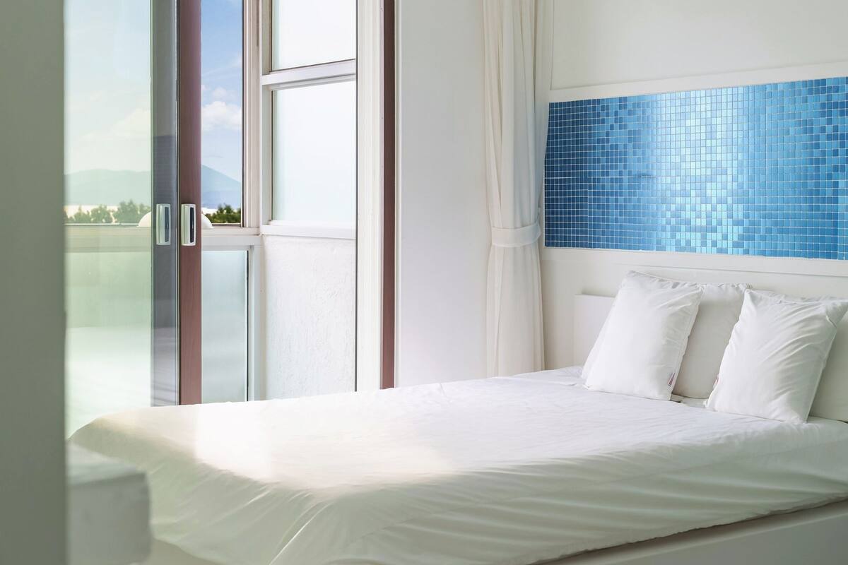 Une chambre à coucher avec un lit, du linge de lit blanc, une vue par la fenêtre et un mur recouvert de carreaux bleus.