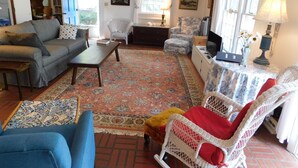 Uma sala de estar acolhedora com um sofá, poltrona e uma mesa pequena. Há um tapete estampado no chão e uma janela com cortinas.