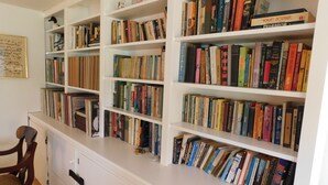Um quarto com uma estante branca repleta de livros, uma cadeira de madeira e uma escrivaninha branca com gavetas.