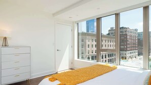 A bedroom with a large window, a bed with a yellow blanket, and a white dresser. - Central 2BR Condo in Downtown Boston & T-Station (Boston)