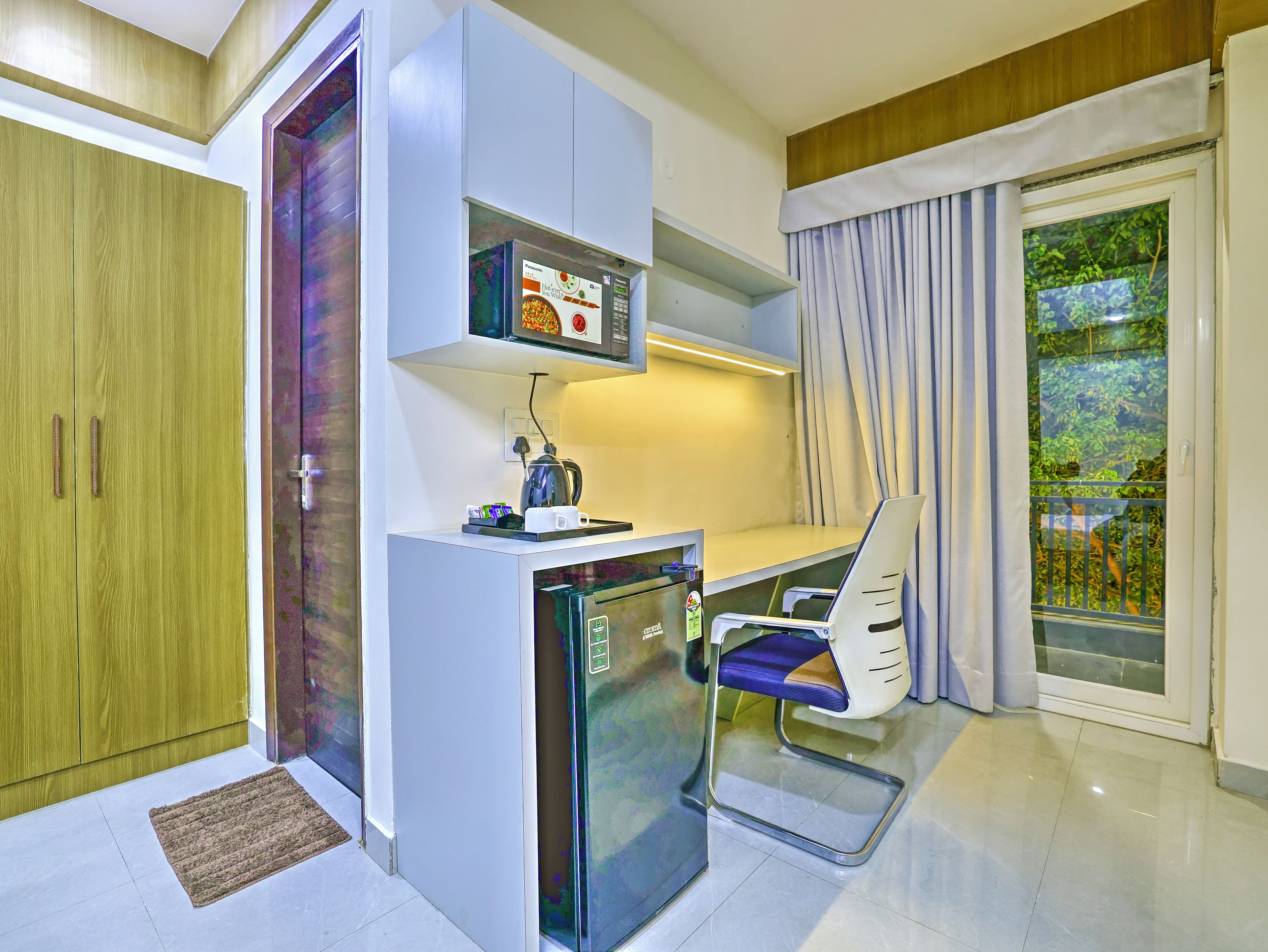 A compact kitchen with a microwave, kettle, and a small refrigerator. There is a chair with a tray table and a window with curtains.