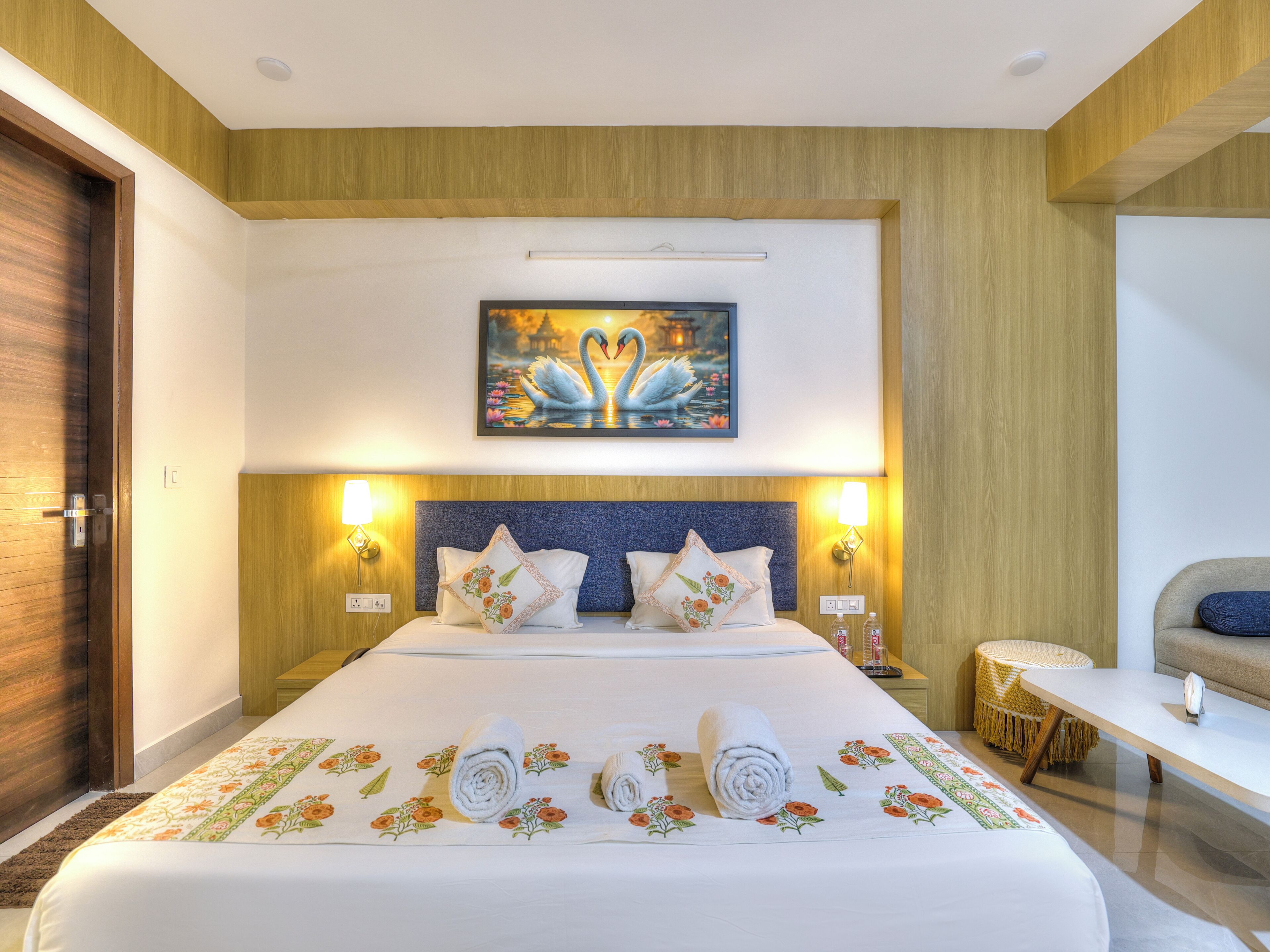 A hotel room with a large bed, two bedside lamps, a painting of swans, and a small round table.