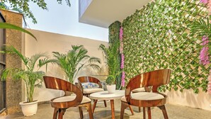 Un patio con dos sillas de madera y una mesita pequeña, rodeado de plantas en macetas y un muro decorativo con plantas trepadoras.