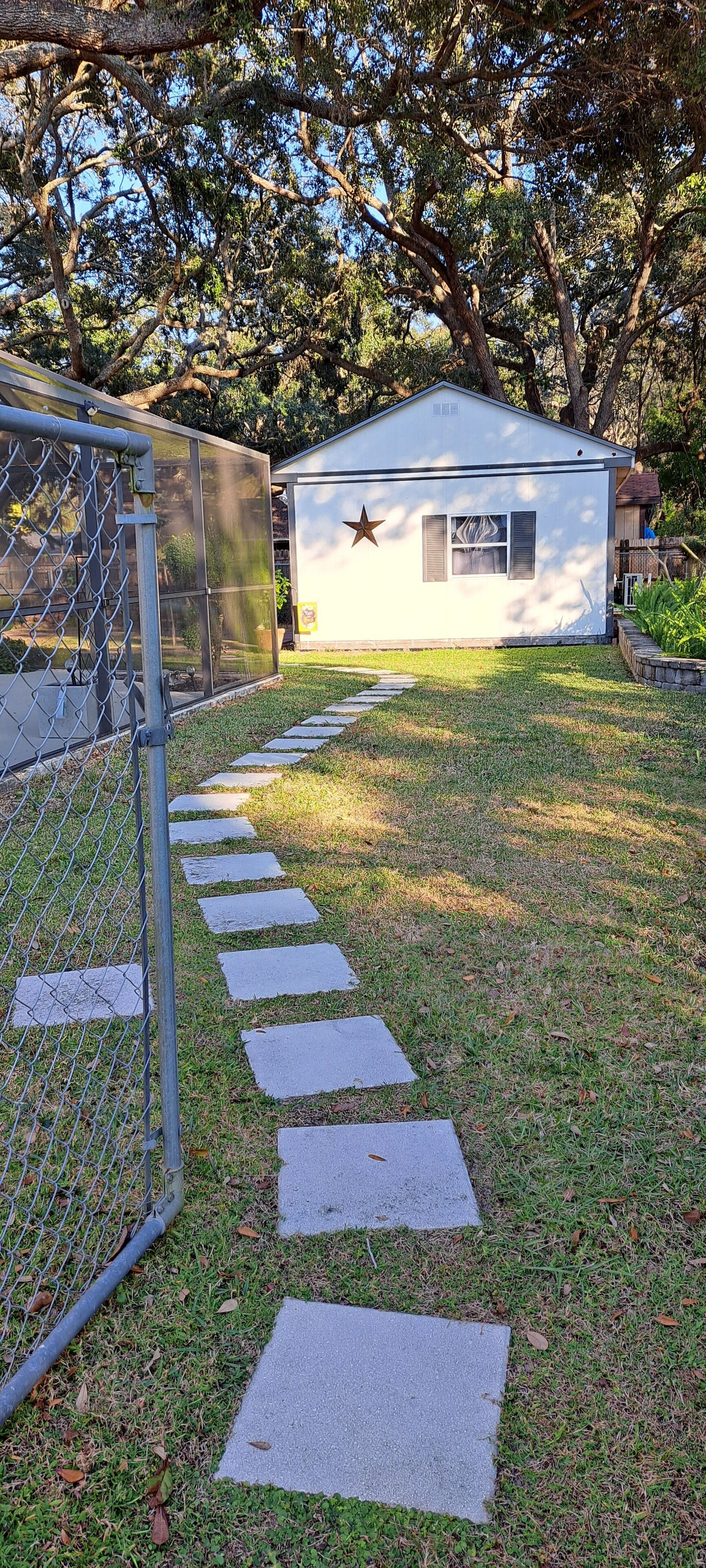 A fenced yard with a pathway leading to a small white building with a star on the door, surrounded by trees.