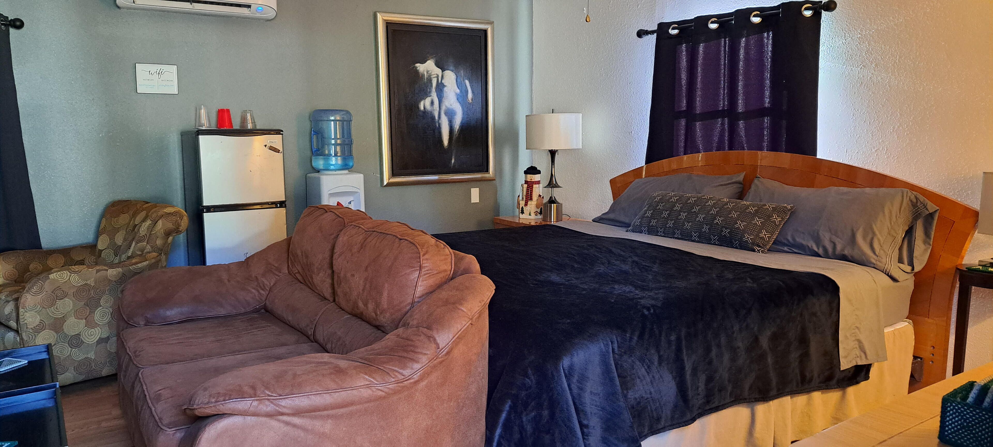 A hotel room with a bed, a couch, a water dispenser, a refrigerator, a lamp, and a painting on the wall.