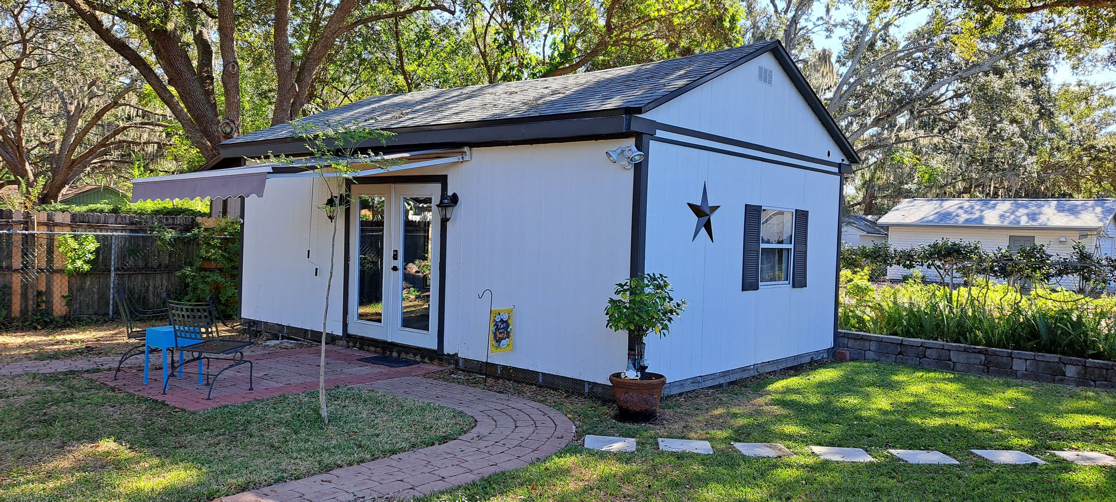 Une petite maison de plain-pied, à l’extérieur blanc et aux bordures noires, dotée d’un patio couvert avec un banc bleu et un pot de fleurs.