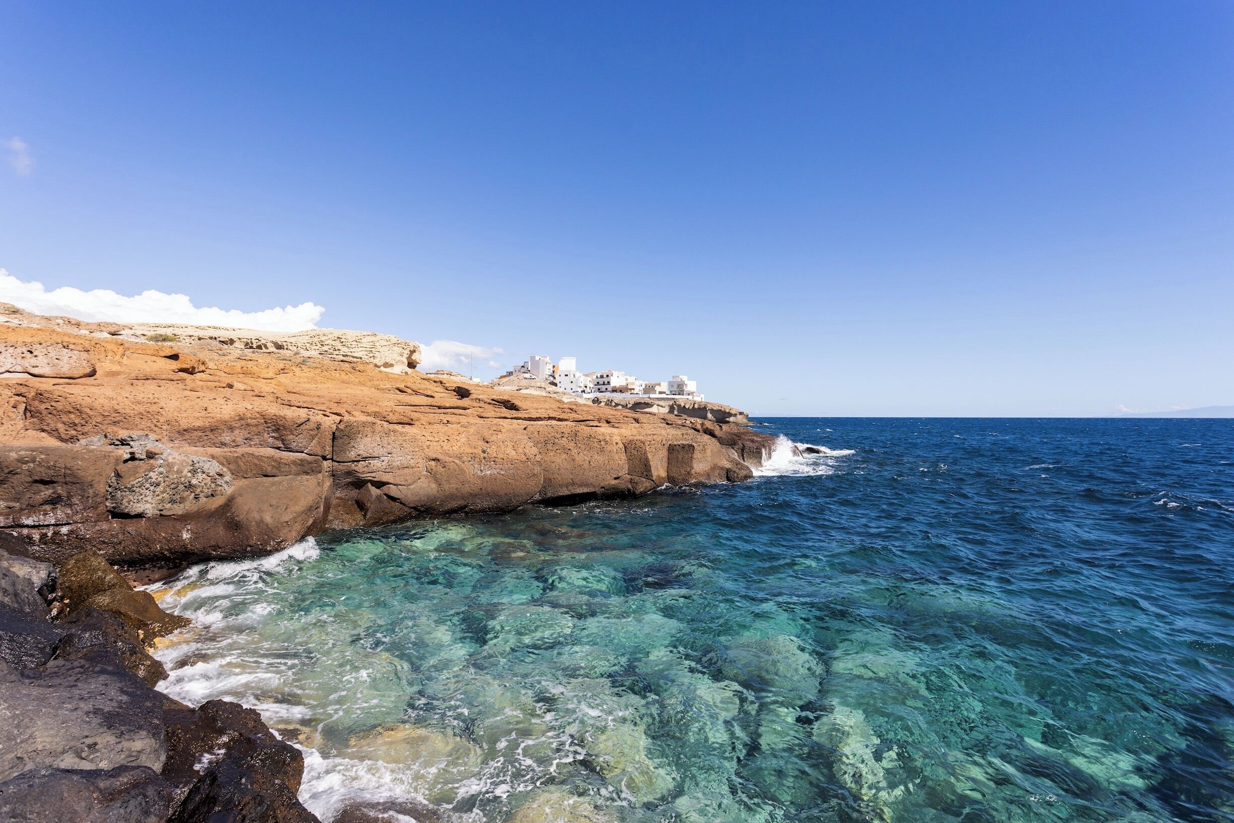 A coastal landscape with clear blue waters, rocky terrain, and white buildings on a cliff.