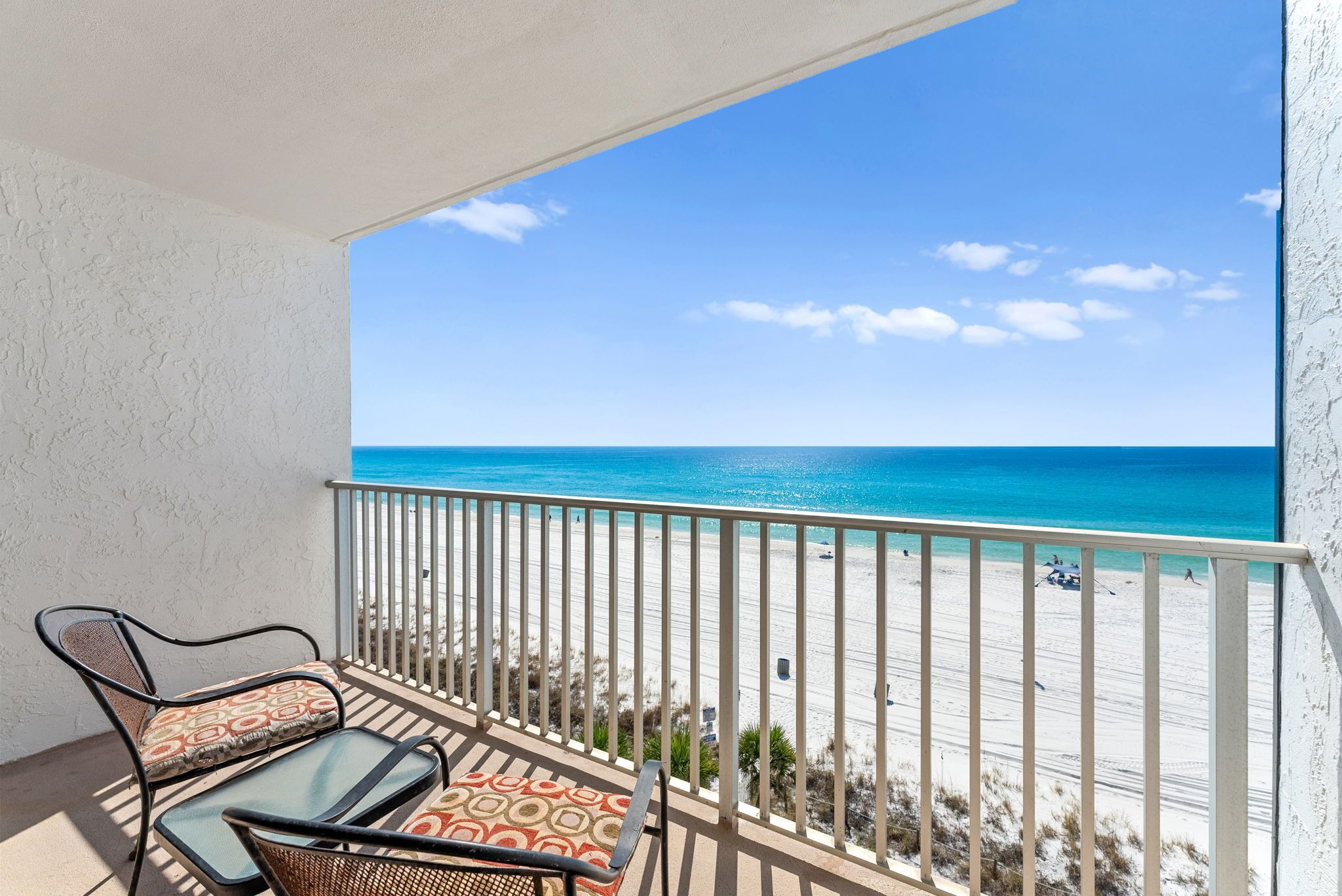 A balcony with two chairs and a table, overlooking a beach with a clear blue sky and ocean.