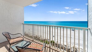 Een balkon met twee stoelen en een tafel, met uitzicht op een strand en een helderblauwe lucht en oceaan.
